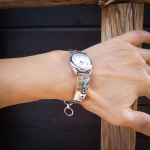 Könnte beinhalten: Eine silberfarbene Armbanduhr mit weißem Zifferblatt und schwarzen Ziffern. Die Uhr hat ein dekoratives silbernes Armband mit offenem Design. Die Uhr befindet sich an einem Handgelenk.