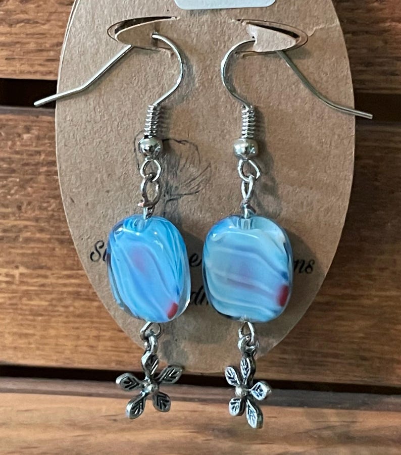 May include: Blue and white glass square dangle earrings with silver flower charms. The earrings have silver hooks.