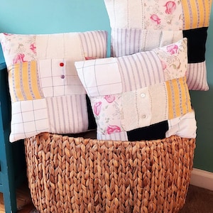 May include: Three square throw pillows featuring a patchwork design of floral, striped, and solid fabric panels in white, yellow, pink, and black. The pillows include small decorative buttons and are displayed inside a large, round, woven brown basket.