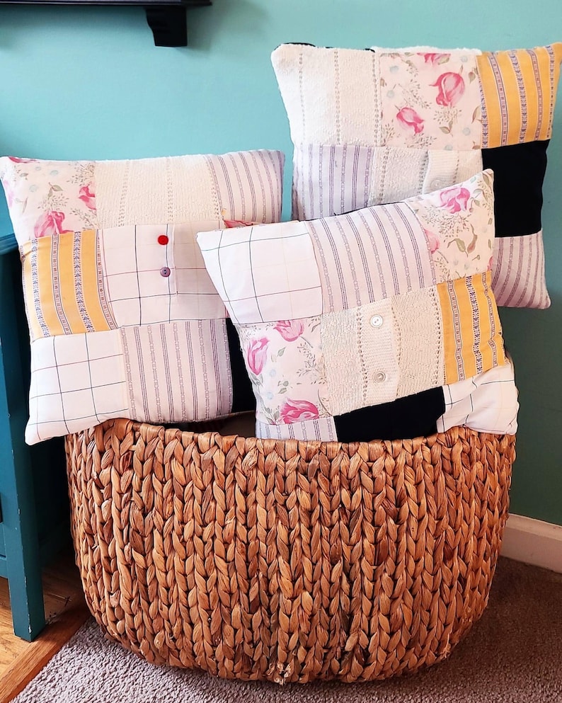 May include: Three square throw pillows featuring a patchwork design of floral, striped, and solid fabric panels in white, yellow, pink, and black. The pillows include small decorative buttons and are displayed inside a large, round, woven brown basket.