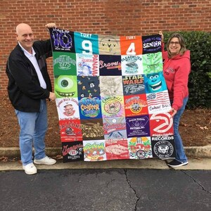May include: A colorful quilt made from various t-shirts, featuring logos, graphics, and text. The quilt is held up by two people. The t-shirts include logos for sports teams, schools, and other organizations. The quilt is a mix of colors.