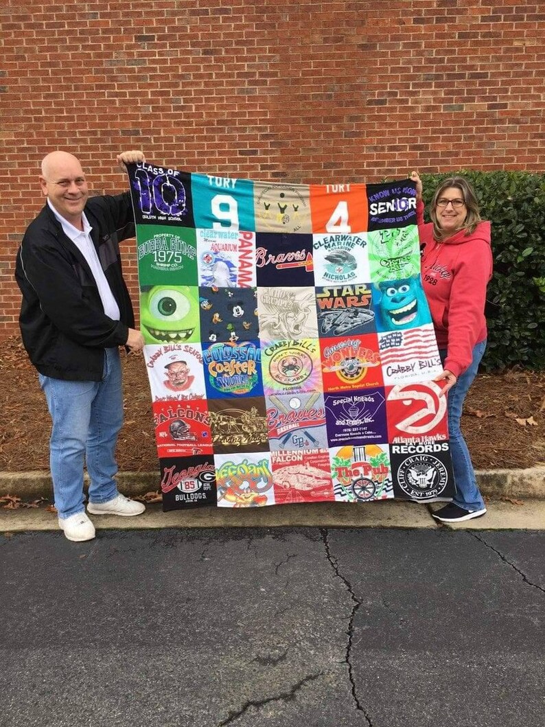 May include: A colorful quilt made from various t-shirts, featuring logos, graphics, and text. The quilt is held up by two people. The t-shirts include logos for sports teams, schools, and other organizations. The quilt is a mix of colors.