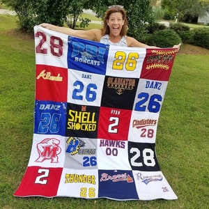 May include: A white, red, blue, and black patchwork quilt made from old sports jerseys. The quilt features various team names and numbers, including "Bobcats", "Gladiator", "Hawks", "Thunder", and "Braves".