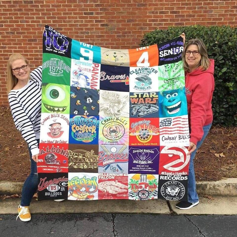 May include: A handmade quilt made from repurposed t-shirts. The quilt features a variety of colorful graphic designs and logos, including sports teams, cartoon characters, and vacation destinations. The quilt is held up by two people.