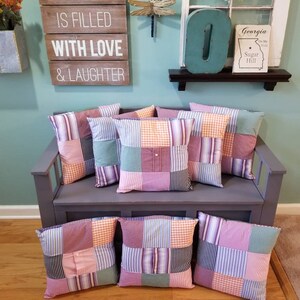 May include: Six patchwork pillows with a variety of colors and patterns, including pink, green, blue, and orange, are arranged on a gray bench. The pillows are made from repurposed fabric and have a rustic, farmhouse style. The bench is in front of a wall with a wooden sign that reads "This home is filled with love & laughter".