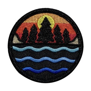 May include: A circular embroidered patch with a sunset scene over a lake. The sunset is in shades of orange, red, and yellow. There are five dark green trees in front of the sunset. The lake is blue and has three waves. The patch is on a black background.