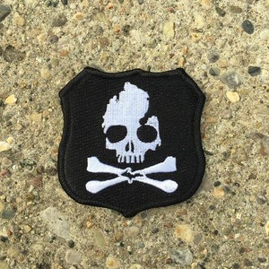 May include: Black and white embroidered patch shaped like a shield with a skull and crossbones. The skull is outlined with the shape of the state of Michigan.