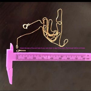 May include: A gold chain with a clasp, measured with a purple ruler. The ruler is marked in inches and millimetres. The text on the ruler reads "Check the size of the end hole of your own chain to see if this charm fits through before purchase."