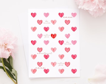 May include: A white greeting card with watercolour hearts in shades of pink and red. The card features phrases like "Be mine" and "XOXO". Pink peonies and a gold geometric candle holder are also in the image.