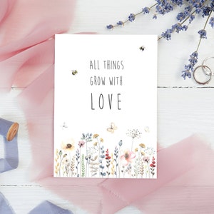 May include: A white card with the text "All things grow with love" in black lettering. The card is decorated with a watercolor illustration of a field of wildflowers and bees.