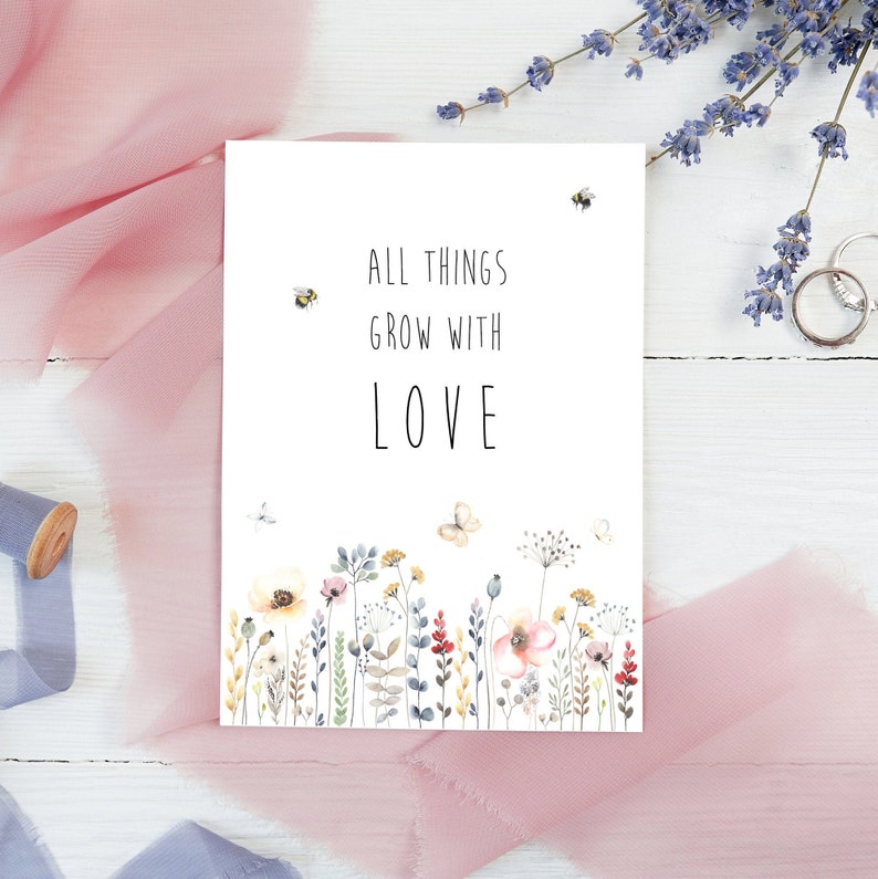 May include: A white card with the text "All things grow with love" in black lettering. The card is decorated with a watercolor illustration of a field of wildflowers and bees.