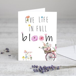 May include: A white card with the text "Live life in full bloom" in black lettering. The card features a watercolor illustration of a bicycle with a basket full of flowers and butterflies.