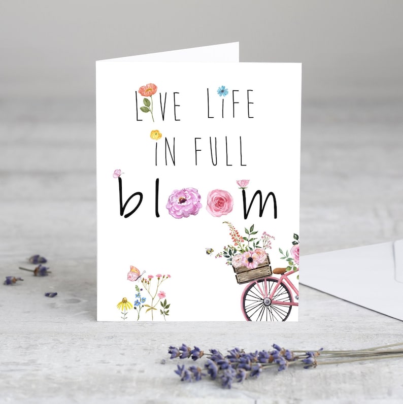 May include: A white card with the text "Live life in full bloom" in black lettering. The card features a watercolor illustration of a bicycle with a basket full of flowers and butterflies.