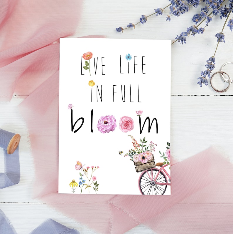 May include: White card with the text "Live Life in Full Bloom" in black lettering. A pink bicycle with a basket full of colorful flowers is in the bottom right corner.