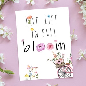 May include: White card with the text "Live Life in Full Bloom" in black lettering. Watercolor flowers and a pink bicycle with a basket full of flowers are illustrated on the card.