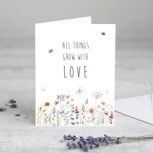 May include: A white card with a watercolor illustration of a field of flowers and bees. The text on the card reads "All things grow with love."