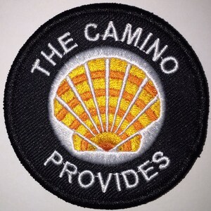 Op de afbeelding: Zwart en wit geborduurde patch met een gele en oranje schelp en de tekst "THE CAMINO PROVIDES".