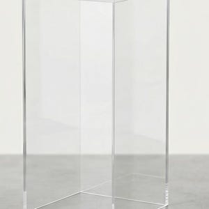May include: A tall, rectangular display pedestal constructed from clear, transparent acrylic. The structure features four vertical sides and a flat top surface, all joined with clean, sharp edges to form a hollow, open-bottomed box shape.