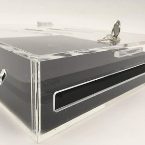 May include: A clear acrylic case with a lock and key, designed to hold and protect a black gaming console. The case has a sleek and modern design.