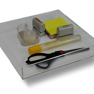 May include: A clear plastic tray with a variety of office supplies, including a tape dispenser, a pair of scissors, a glue stick, a yellow sticky note, and two small office punches labeled "DRAFT" and "FAX TO".