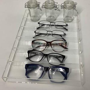 May include: A clear acrylic display stand holding five pairs of eyeglasses and three small glass jars with metal clasps. The eyeglasses have various frame styles and colors, including black, brown, and blue. The jars are empty.