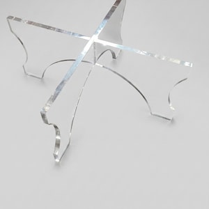 May include: A clear acrylic display stand with a four-way cross design. Each of the four arms has a curved edge and a decorative cutout. The stand is designed to hold an object.