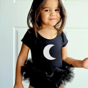 Puede incluir: Una niña pequeña con un leotardo negro con un diseño de media luna blanca y un tutú de tul negro.