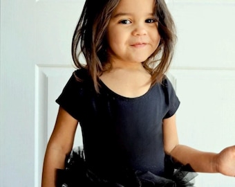 May include: A black short-sleeved leotard featuring a layered tulle tutu skirt attached at the waist, paired with a headband adorned with black and white cat ears. The garment is solid black with a round neckline and a full, ruffled tulle skirt.