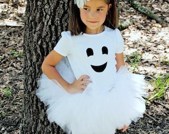 May include: A child's Halloween costume featuring a white t-shirt with a black ghost face, a white tutu skirt, and white leggings. A white flower hair accessory completes the look. The child is standing next to a tree.