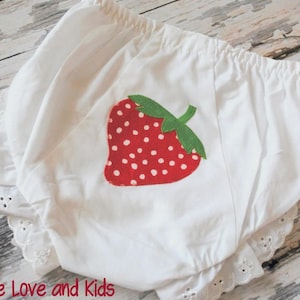 May include: White baby bloomers with a red strawberry applique. The strawberry has white seeds and a green stem. The bloomers have a ruffled eyelet lace trim and an elastic waistband. The text "Peace Love and Kids" is at the bottom.