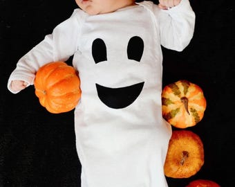 May include: A white baby sleeper with a black ghost face on the front. The sleeper is surrounded by small orange pumpkins.