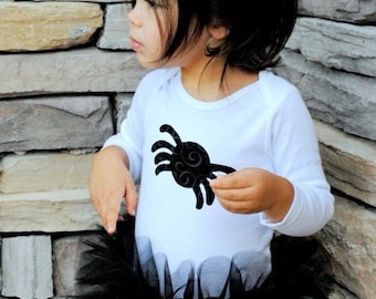 Könnte beinhalten: Ein kleines Kind trägt ein weißes Langarmshirt mit einem schwarzen Spinnenmotiv und einen schwarzen Tüll-Tutu-Rock.