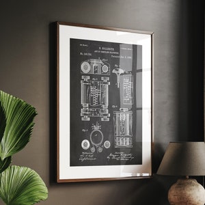 May include: A black and white vintage patent drawing of a "Hydraulic Refrigerant" by E. Bollente, patented in 1898. The drawing is framed and hanging on a dark wall above a wooden desk with a lamp and books.