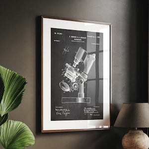 Può includere: Un disegno di brevetto vintage in bianco e nero di un microscopio, incorniciato in una cornice di legno scuro, appeso a una parete scura sopra una scrivania di legno con una lampada e dei libri.