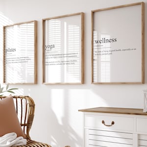 May include: A wicker chair with a brown throw blanket and a pillow sits in a room with three framed prints. The prints have the words "pilates", "yoga", and "wellness" with their definitions. A white cabinet with two drawers and a wooden top is in the background.