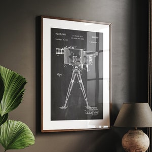 Puede incluir: Un dibujo de patente vintage en blanco y negro de una cámara en un trípode. El número de patente es 2.883.537 y la fecha es el 23 de septiembre de 1959. El dibujo está enmarcado en un marco de madera marrón y colgado en una pared oscura.