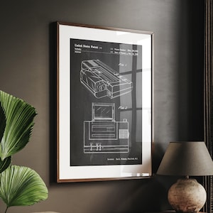 Könnte beinhalten: Eine Schwarzweiß-Patentzeichnung einer Vintage-Videospielkonsole. Die Zeichnung zeigt die Ansicht von oben und die Seitenansicht der Konsole. Die Patentnummer und das Datum sind oben auf der Zeichnung aufgeführt. Die Zeichnung ist in einem braunen Holzrahmen gerahmt und hängt an einer dunklen Wand.