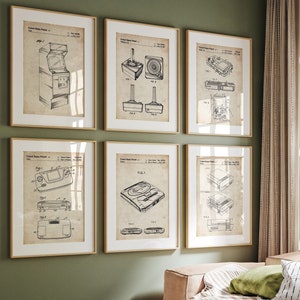 Op de afbeelding: Een set van vijf vintage-stijl prints met zwart-wit illustraties van klassieke videogameconsoles. De prints zijn ingelijst in een licht houten lijst en worden weergegeven op een groene muur.
