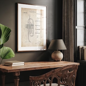 Op de afbeelding: Een ingelijste vintage print van een trompet patent illustratie. Het kunstwerk toont gedetailleerde technische tekeningen in sepia tinten, tegen een witte achtergrond, en is gevat in een bruine houten lijst. De print wordt getoond op een houten bureau met een lamp en een decoratieve stoel.