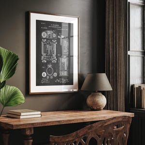 May include: A framed black and white print of a vintage patent diagram. The diagram is of a mechanical device, with detailed illustrations and technical annotations. The frame is a warm brown, and the print is displayed on a dark wall, with a wooden desk in the foreground.