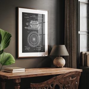 May include: A framed black and white print of a Tesla Dynamo Electric Machine patent diagram. The print is in a brown frame with a white mount. The print is displayed on a wooden desk with a lamp and books.