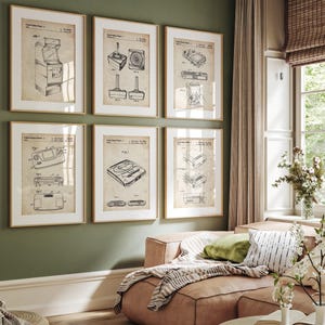 May include: Six framed vintage video game patent prints hang on a green wall. The prints feature detailed technical drawings of classic gaming consoles and controllers, with a neutral color palette and gold frames. The room includes a pink sofa and decorative pillows.