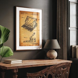 May include: Framed vintage patent print of a laptop computer. The print features detailed technical drawings in black ink on a sepia-toned background, with the United States Patent information at the top. The print is displayed in a brown wooden frame.