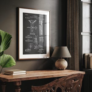 May include: A framed patent print of an apparatus for electric welding, with a dark background and white diagrams. The print is in a brown frame and is displayed on a dark wall above a wooden desk. A lamp with a gray shade sits on the desk.