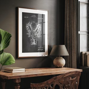 May include: Framed black and white print of an engine diagram. The print is in a brown frame with a white mount. The diagram is detailed and technical, with various components labelled. The print is displayed on a dark wall above a wooden desk.