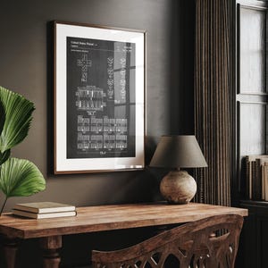 May include: A framed black and white print of a United States patent for a game. The print is in a brown frame and is displayed on a wooden desk. A lamp with a grey shade sits on the desk, and a green plant is visible.