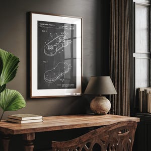 May include: Framed patent print of a video game controller in black and white. The print features two detailed diagrams labeled FIG. 1 and FIG. 2, with technical annotations. The frame is a warm brown, and the print is displayed on a dark wall.