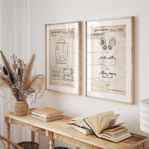 Puede incluir: Dos impresiones enmarcadas de estilo vintage con diseños de mandos de videojuegos. Las impresiones están en marcos de madera y presentan diagramas detallados sobre un fondo beige. Se exhiben sobre una consola de madera con libros y un jarrón decorativo.