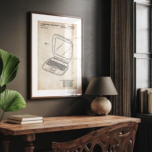 May include: Framed vintage laptop patent print. The artwork features a detailed blueprint of a laptop computer, with technical drawings and annotations. The print is in a wooden frame, displayed on a wooden desk with a lamp and books.