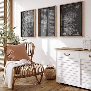 Op de afbeelding: Drie ingelijste blauwdrukken van muziekinstrumenten hangen aan een witte muur. De prints, in houten frames, tonen gedetailleerde diagrammen van een trompet, een Franse hoorn en een ander instrument. Een rieten stoel met een kussen en deken staat eronder.